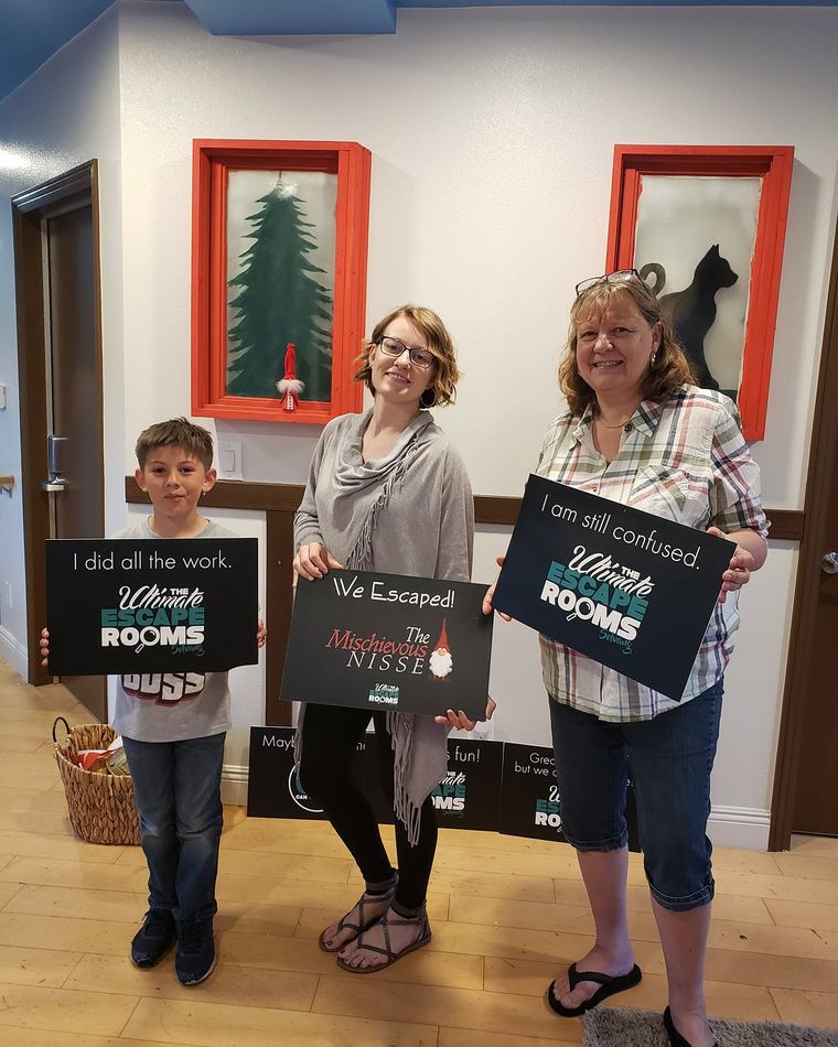 A boy and two women in the hallway at The Ultimate Escape Rooms Solvang holding signs that read "I did all the work.", "We Escaped!" and "I am still confused."