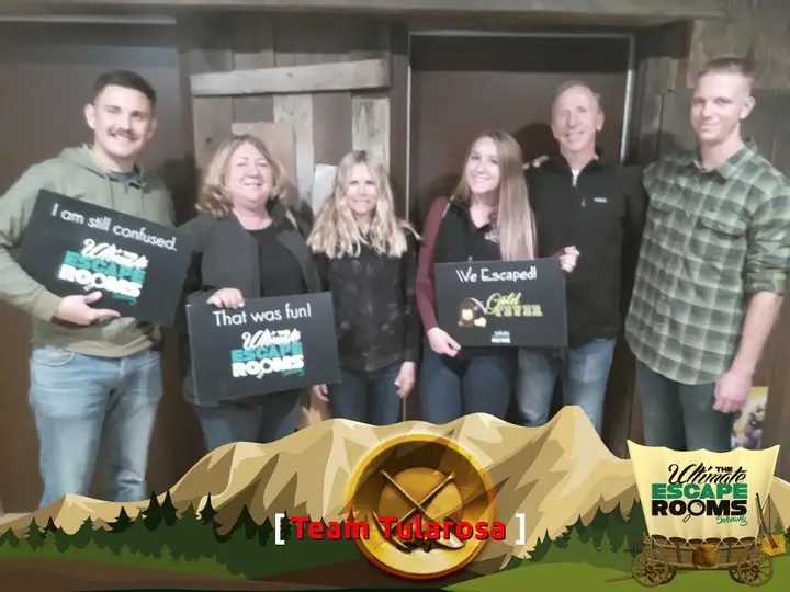 A group of six at The Ultimate Escape Rooms Solvang holding signs that read "I am still confused.", "That was fun!" and "We Escaped!" in front of the Gold Fever door